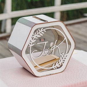 May include: White hexagonal wooden box with a clear acrylic front panel. The front panel features a laser-cut design of two initials, "J" and "K", intertwined with a heart and floral accents. The text "2 & 1" is visible in the background.