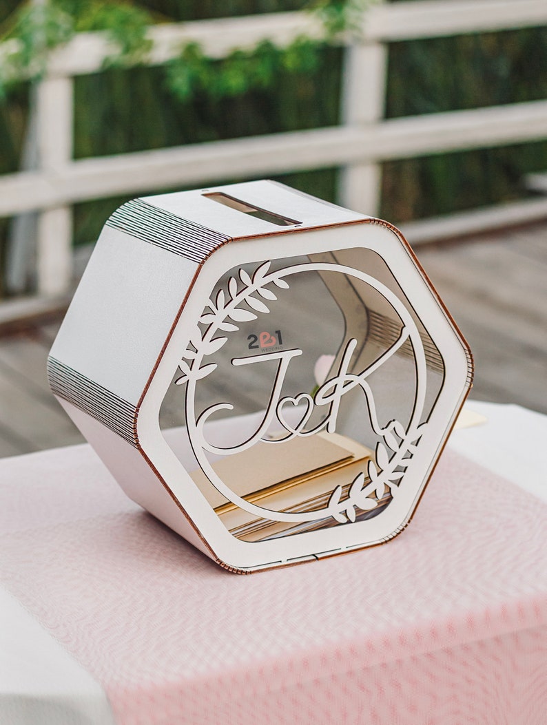 May include: White hexagonal wooden box with a clear acrylic front panel. The front panel features a laser-cut design of two initials, "J" and "K", intertwined with a heart and floral accents. The text "2 & 1" is visible in the background.