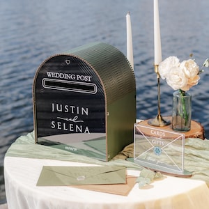 May include: A decorative wedding post box with the words "Wedding Post" and the names "Justin and Selena" in white script. The box is olive green and sits on a table with a white tablecloth. A sign that says "Cards and Gifts" is also on the table.