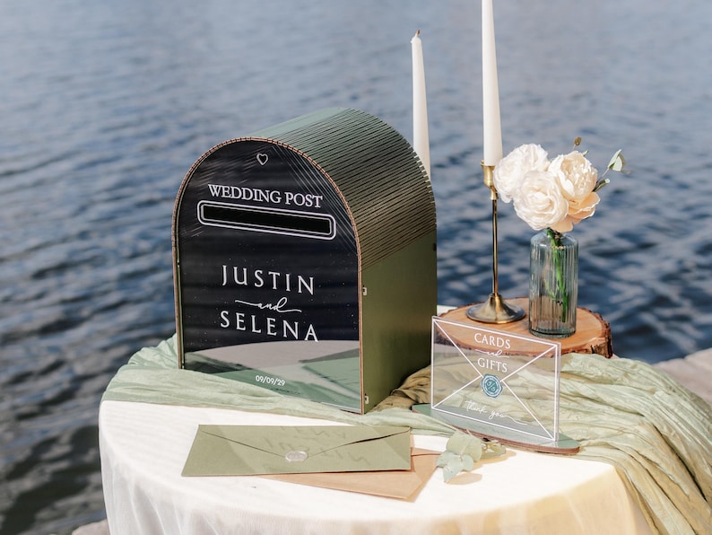 May include: A decorative wedding post box with the words "Wedding Post" and the names "Justin and Selena" in white script. The box is olive green and sits on a table with a white tablecloth. A sign that says "Cards and Gifts" is also on the table.