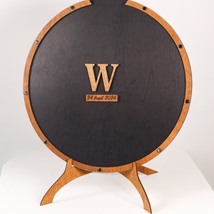 May include: A round wooden sign with a black center and a wooden stand. The sign has a large wooden letter "W" and the date "24 August 2024" written on it.