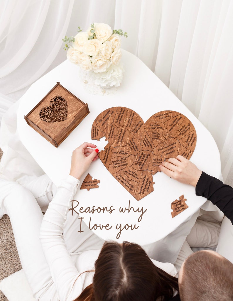 Puede incluir: Un rompecabezas de madera con forma de coraz&oacute;n con razones grabadas por las que alguien podr&iacute;a amar a otra persona. El rompecabezas se est&aacute; ensamblando en una mesa blanca con una caja de madera y un ramo de flores blancas en el fondo. El texto "Reasons why I love you" est&aacute; escrito debajo del rompecabezas.