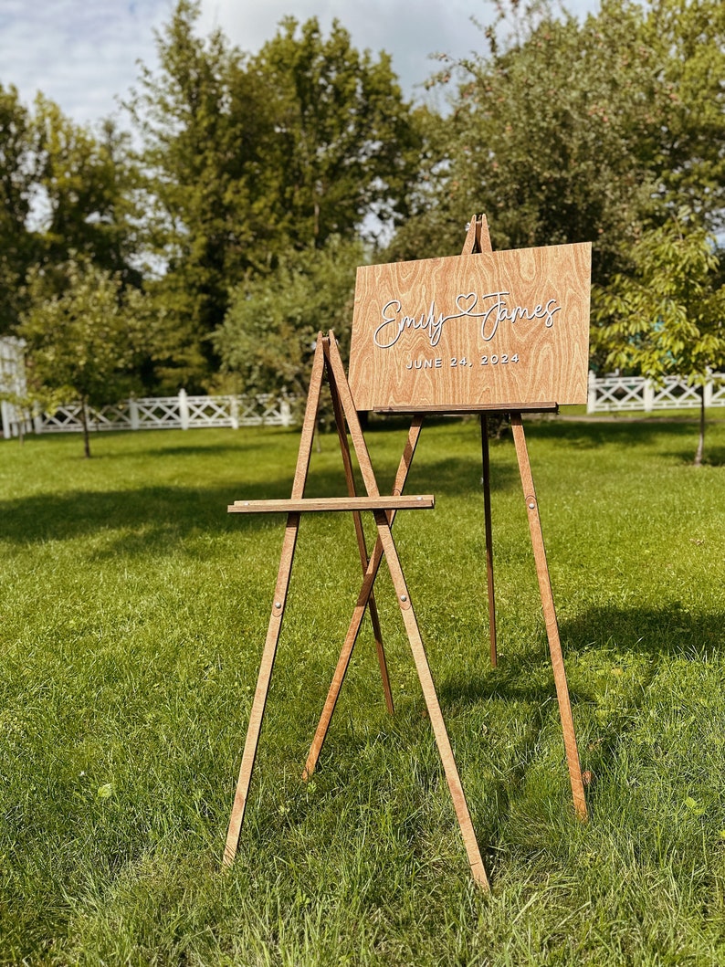 May include: A wooden easel with a sign that reads "Emily & James June 24, 2024" in white script. The sign is on a wooden background with a wood grain pattern.