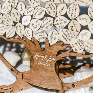 Puede incluir: Un libro de visitas de madera con forma de &aacute;rbol con hojas para que los invitados escriban mensajes. El &aacute;rbol est&aacute; decorado con flores blancas y el texto "Henry & Laura 12/12/24" est&aacute; escrito en el tronco.