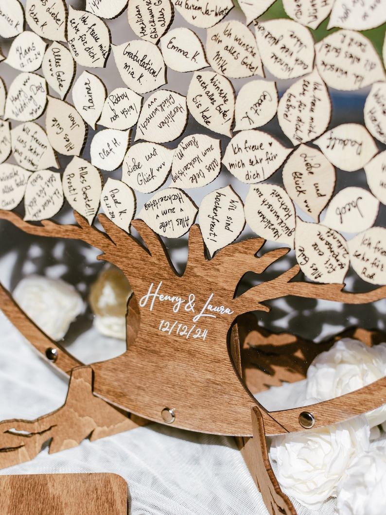 Puede incluir: Un libro de visitas de madera con forma de &aacute;rbol con hojas para que los invitados escriban mensajes. El &aacute;rbol est&aacute; decorado con flores blancas y el texto "Henry & Laura 12/12/24" est&aacute; escrito en el tronco.