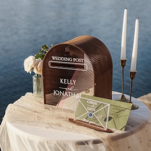May include: A wooden wedding post box with the words "WEDDING POST" and the names "KELLY and JONATHAN" printed on the front. Two white candles in brass holders and a "CARDS and GIFTS" sign are also on the table. A vase of white flowers sits to the left.