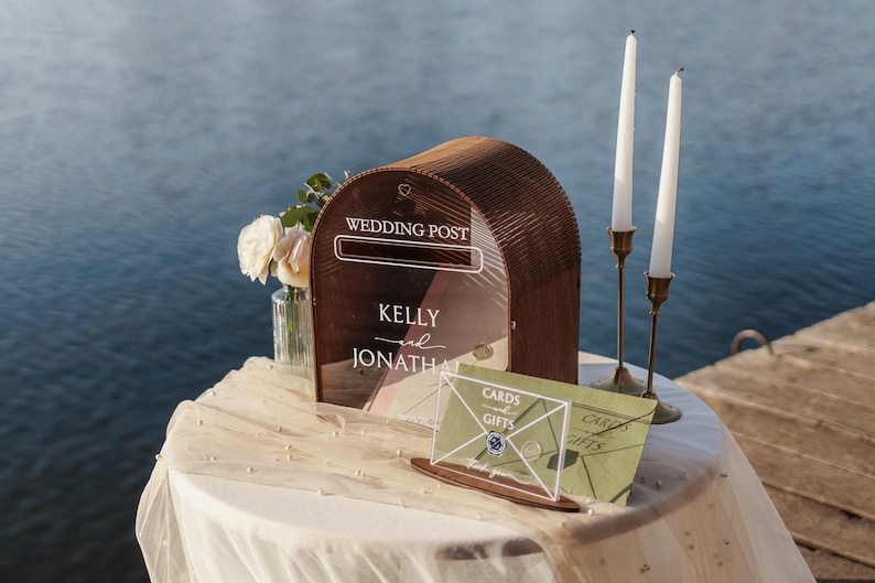 May include: A wooden wedding post box with the words "WEDDING POST" and the names "KELLY and JONATHAN" printed on the front. Two white candles in brass holders and a "CARDS and GIFTS" sign are also on the table. A vase of white flowers sits to the left.
