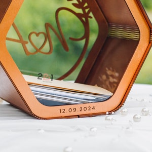 May include: A wooden box with a clear window, featuring a stylized design of two initials with a heart and a leafy branch. The box has a date engraved on the bottom: 12.09.2024. The text "2021 Wedding" is also visible on the box.
