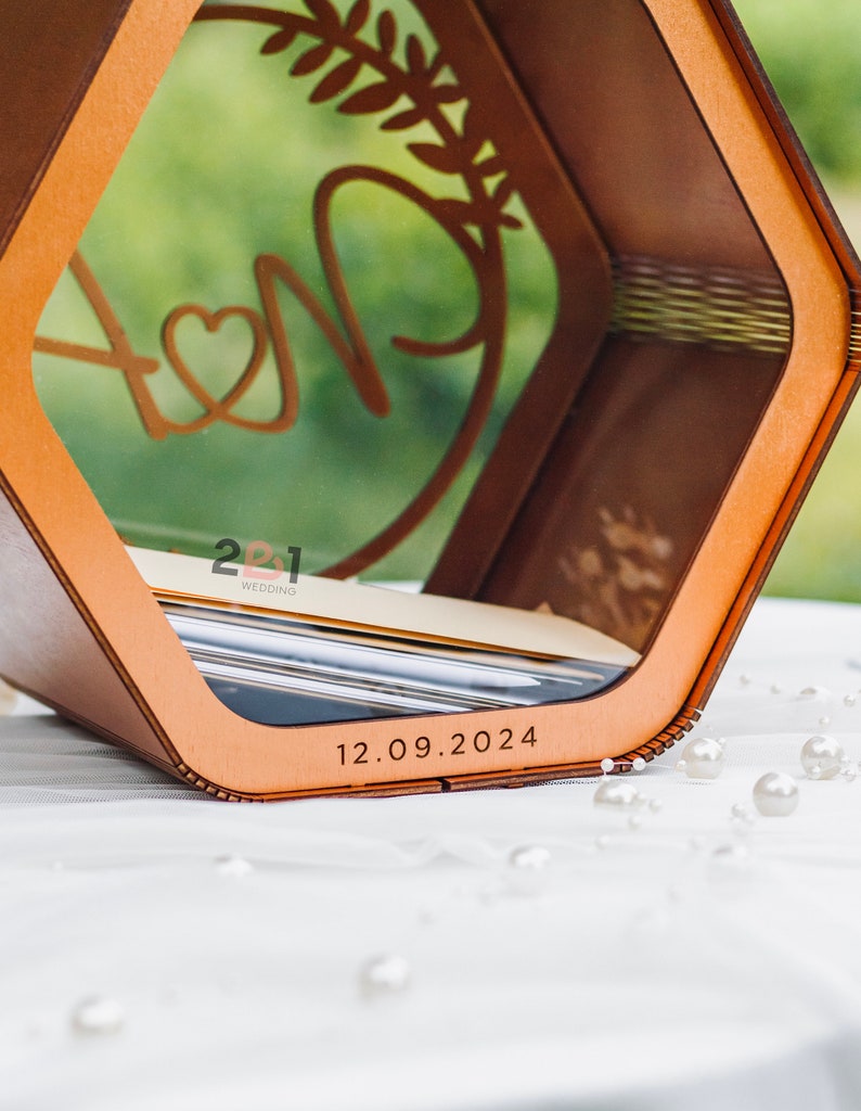 May include: A wooden box with a clear window, featuring a stylized design of two initials with a heart and a leafy branch. The box has a date engraved on the bottom: 12.09.2024. The text "2021 Wedding" is also visible on the box.