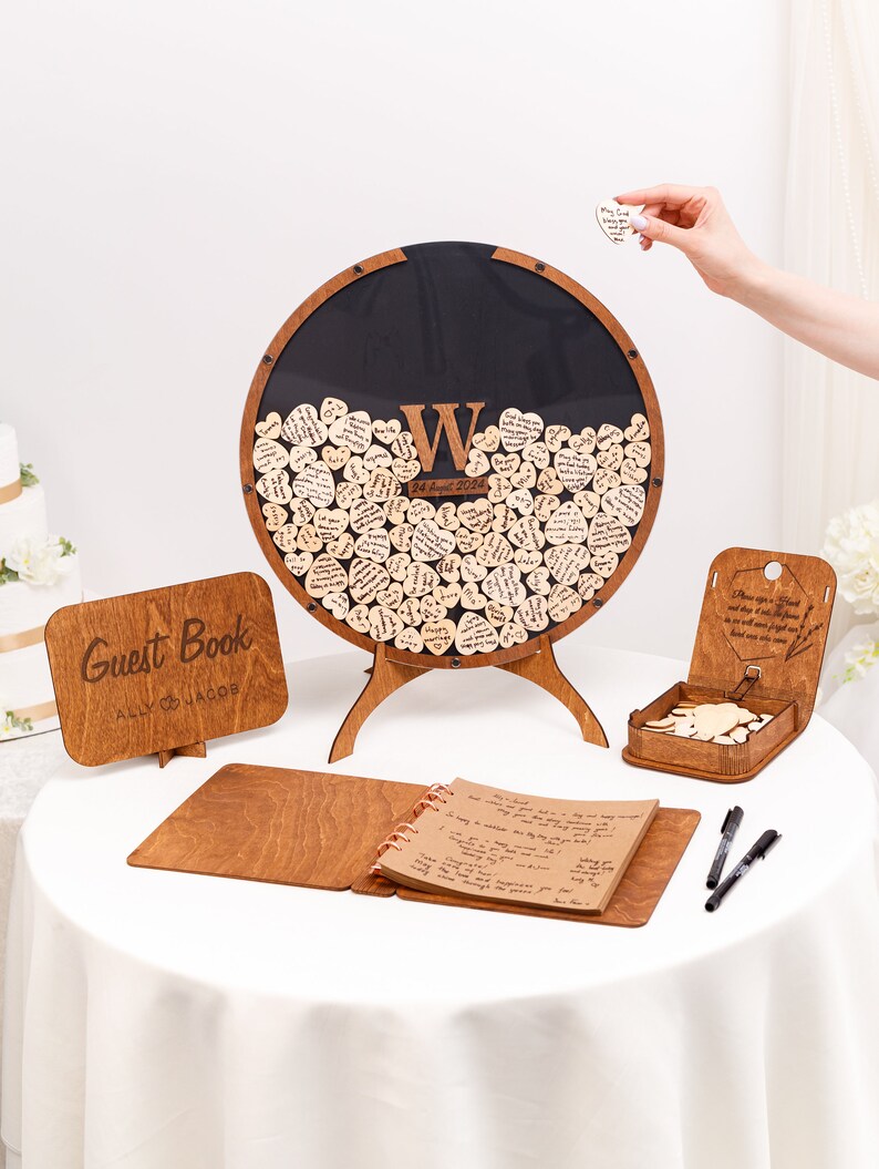 May include: A wooden guest book set with a heart-shaped wooden guest book, a wooden sign that says "Guest Book Ally & Jacob", a wooden guest book with a brown cover, and two black pens.