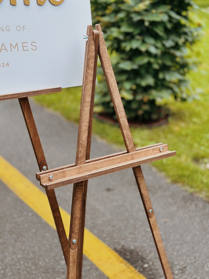 May include: A wooden easel with a white sign that reads "Wedding of James 2024". The easel is standing on a paved surface with a yellow line.