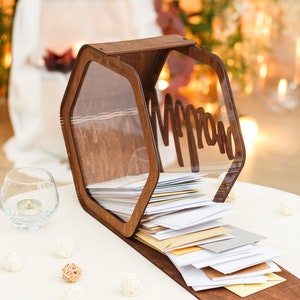 May include: A wooden hexagonal box with a clear acrylic lid, labeled "Mr. & Mrs." and filled with white envelopes. The box is sitting on a white table with small white and brown decorative balls.