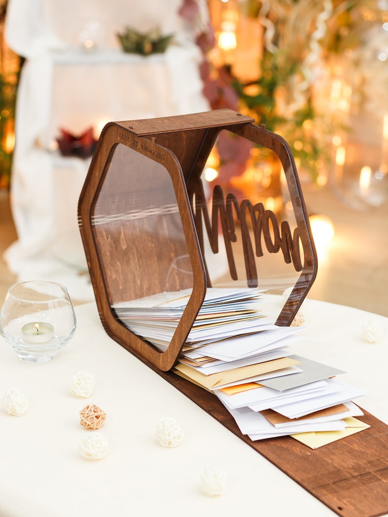May include: A wooden hexagonal box with a clear acrylic lid, labeled "Mr. & Mrs." and filled with white envelopes. The box is sitting on a white table with small white and brown decorative balls.