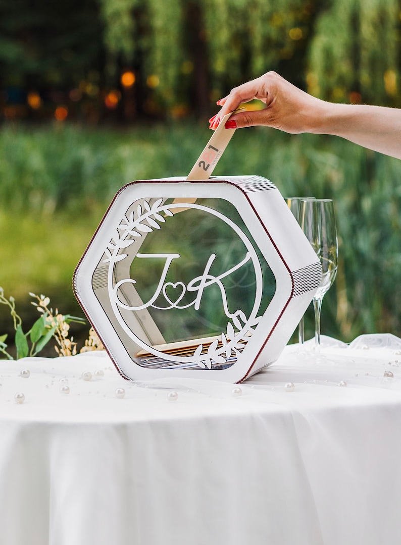 May include: White hexagonal wedding card box with a clear glass insert. The box has a cutout design of leaves and the initials "J & K" with a heart. A hand is placing a card with the number "21" into the box.
