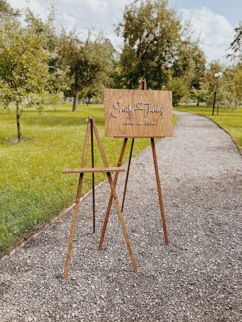 May include: Wooden easel with a sign that reads "Emily & James June 24, 2024". The sign is on a gravel path with green grass and trees in the background.