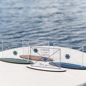 May include: Clear acrylic card and gift signs with a navy blue and teal base. The signs have the words "Cards and Gifts" and "thank you" printed on them. The signs are on a white tablecloth with a water background.