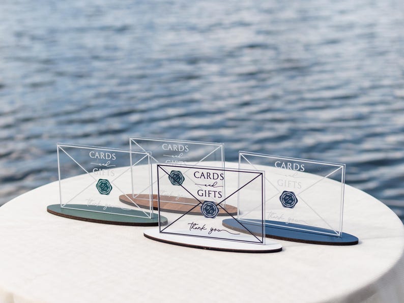 May include: Clear acrylic card and gift signs with a navy blue and teal base. The signs have the words "Cards and Gifts" and "thank you" printed on them. The signs are on a white tablecloth with a water background.