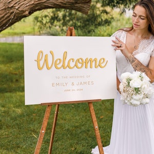 May include: White wedding welcome sign with gold lettering on an easel. The sign reads "Welcome to the wedding of Emily & James June 24, 2024".
