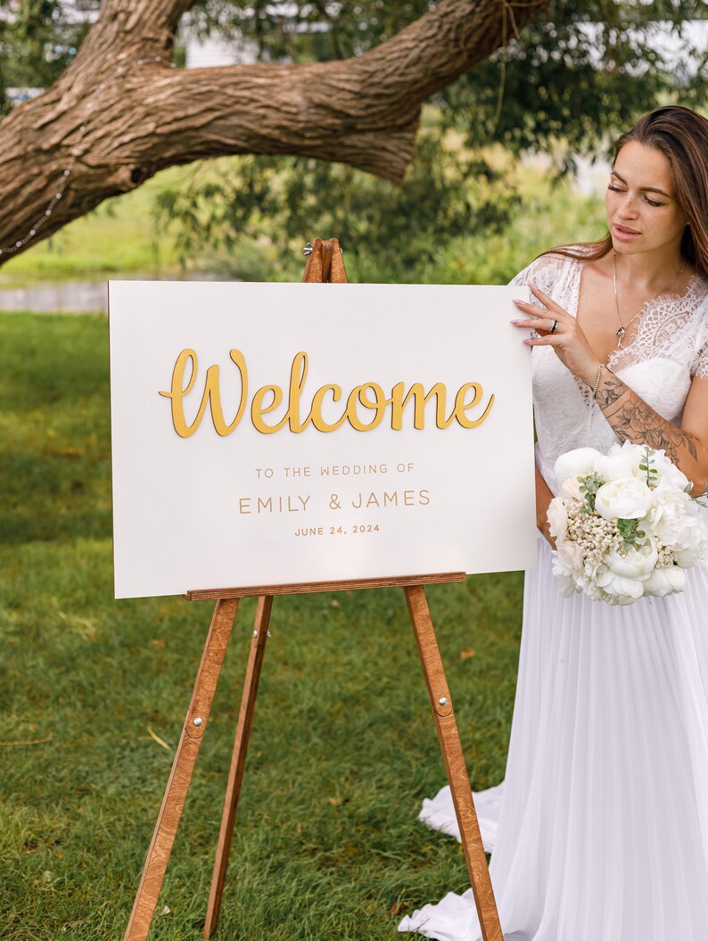 May include: White wedding welcome sign with gold lettering on an easel. The sign reads "Welcome to the wedding of Emily & James June 24, 2024".