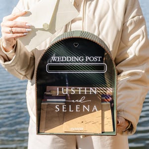 May include: A decorative wedding post box with a black front and a green frame. The box has the words "WEDDING POST" at the top and the names "JUSTIN and SELENA" below. The date "09/09/20" is also visible. An envelope is being placed into the box.