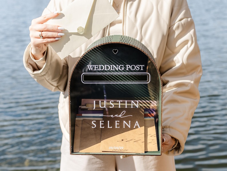 May include: A decorative wedding post box with a black front and a green frame. The box has the words "WEDDING POST" at the top and the names "JUSTIN and SELENA" below. The date "09/09/20" is also visible. An envelope is being placed into the box.