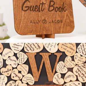 May include: A wooden guest book with the words "Guest Book" and "ALLY JACOB" engraved on it. The book is decorated with a wooden letter "W" and a date "24 August 2024". The letter "W" is surrounded by small wooden hearts with handwritten messages.