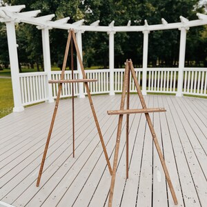 May include: Two wooden easels stand on a light gray wooden deck. The easels are made of natural wood and have a simple design. They are both standing upright and are ready to be used.