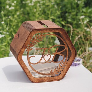 May include: A hexagonal wooden box with a slot on top. The box has a clear acrylic window with a cutout design of two letters, "J" and "B", with a heart in between. The letters are surrounded by a leafy design.