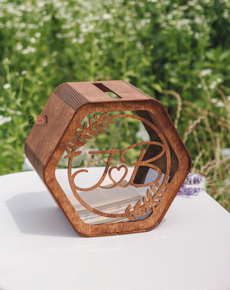 May include: A hexagonal wooden box with a slot on top. The box has a clear acrylic window with a cutout design of two letters, "J" and "B", with a heart in between. The letters are surrounded by a leafy design.