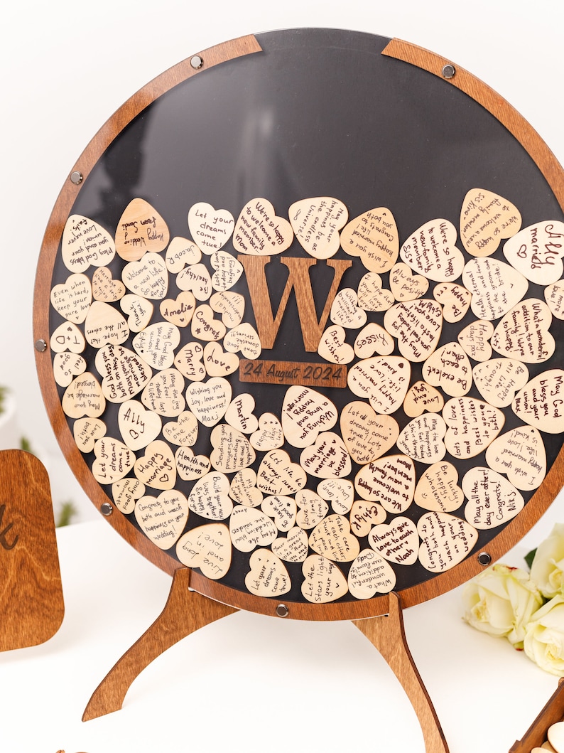 May include: A wooden heart-shaped guest book with a black chalkboard background. The hearts are filled with handwritten messages and the date "24 August 2024" is written on the chalkboard.
