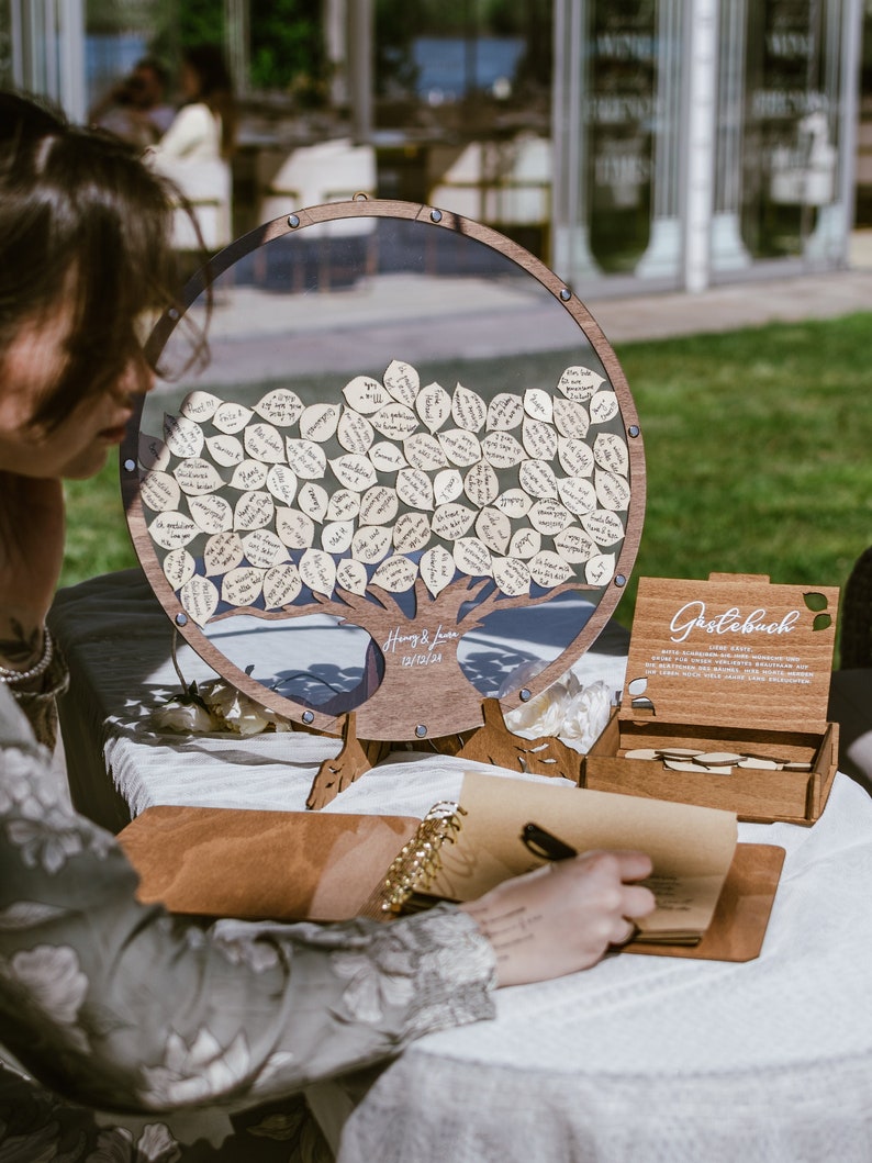 Puede incluir: Un &aacute;rbol de libro de visitas de madera con un marco de acr&iacute;lico transparente lleno de peque&ntilde;as hojas de papel. Las hojas est&aacute;n inscritas con mensajes de los invitados. El &aacute;rbol est&aacute; sobre una mesa con un libro de visitas de cuero marr&oacute;n y un bol&iacute;grafo.