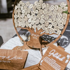 Puede incluir: Un &aacute;rbol de libro de visitas de madera con un c&iacute;rculo de acr&iacute;lico transparente alrededor. El &aacute;rbol est&aacute; decorado con peque&ntilde;as hojas de madera en las que los invitados pueden escribir mensajes. El &aacute;rbol est&aacute; sobre una mesa con una caja de madera y un letrero de madera que dice "Libro de visitas".