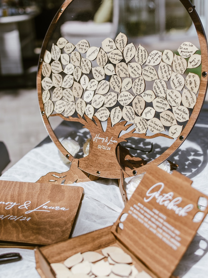 Puede incluir: Un &aacute;rbol de libro de visitas de madera con un c&iacute;rculo de acr&iacute;lico transparente alrededor. El &aacute;rbol est&aacute; decorado con peque&ntilde;as hojas de madera en las que los invitados pueden escribir mensajes. El &aacute;rbol est&aacute; sobre una mesa con una caja de madera y un letrero de madera que dice "Libro de visitas".