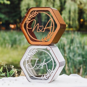 May include: Two hexagonal wooden boxes with clear glass fronts. The top box is brown and features the initials "N & A" with a heart in between. The bottom box is white and features the initials "J & K" with a heart in between. Both boxes have a floral design around the initials.