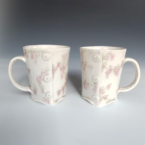 May include: Two white ceramic mugs with a floral pattern in pink and green. The mugs have a unique design with a buttoned front and a curved handle. The mugs are displayed on a light gray surface.