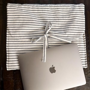 May include: A gray laptop with the Apple logo rests on a black and white striped laptop sleeve. The sleeve is tied with a white ribbon with black stripes. The sleeve is made of fabric.