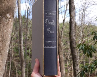 May include: A hardcover book titled "Vanity Fair" by Thackeray, with a dark blue spine and gold lettering. The book is in a light gray slipcase. The image is set against a backdrop of trees.