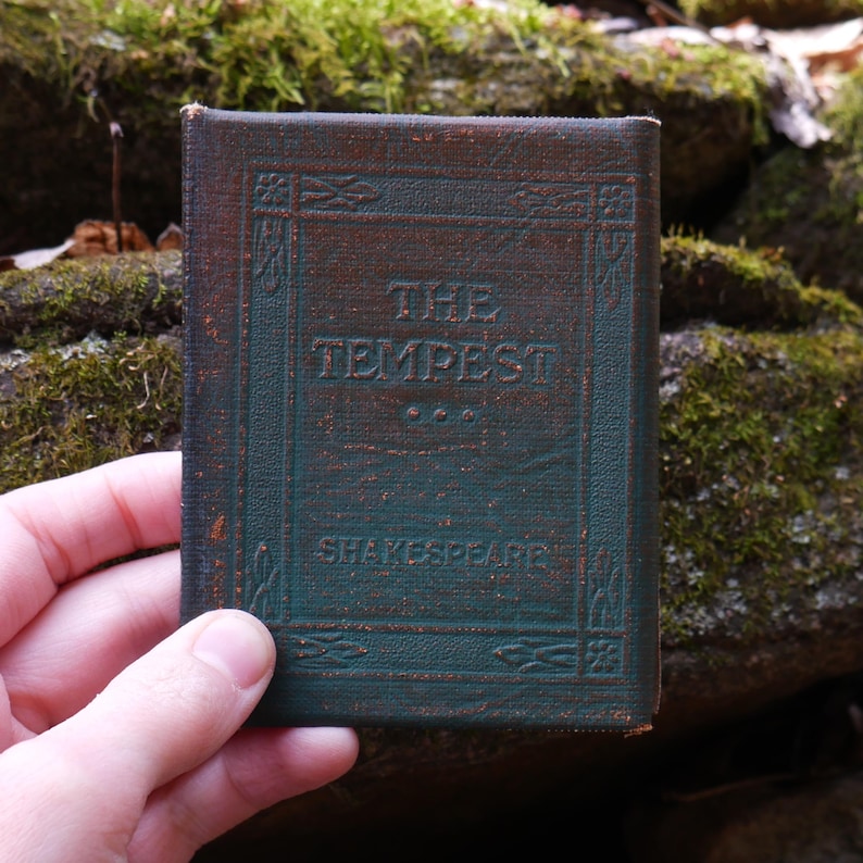 May include: A small, antique book titled "The Tempest" by Shakespeare. The cover is dark green with embossed lettering and decorative borders. The book is held in a hand, with a mossy background.