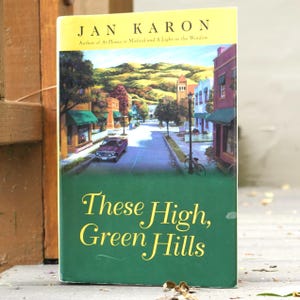 May include: A hardcover book titled "These High, Green Hills" by Jan Karon. The cover features a vibrant illustration of a small town street with shops, a car, and a bicycle. The book's title is in large, elegant script.