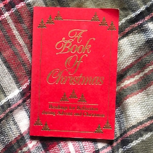 Peut inclure: Un livre rouge intitulé "A Book Of Christmas" avec des lettres dorées et des illustrations de sapins de Noël. La couverture comprend également le texte "Readings for Reflection during Advent and Christmas".
