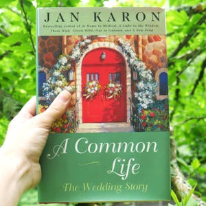 May include: A book titled "A Common Life: The Wedding Story" by Jan Karon. The cover features a painting of a red door with floral arrangements, set against a stone building. The book is held against a backdrop of green foliage.