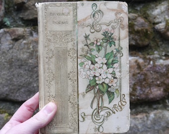 May include: Antique book titled "Bryan's Poems" with a floral design on the cover. The cover is off-white with a faded floral illustration of white flowers and green leaves. The spine has embossed vertical lines and decorative elements. The book is held by a hand.