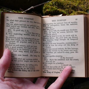 May include: An open, antique book with aged, yellowed pages. The text on the pages is from Shakespeare's "The Tempest." The book is held open by a hand, with the pages showing text and numbers. The book has a brown leather cover.