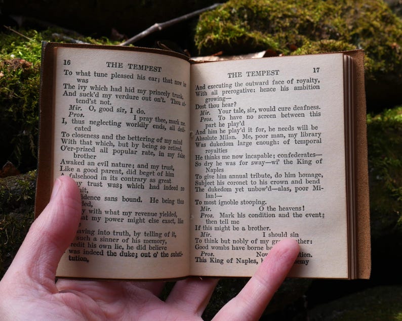 May include: An open, antique book with aged, yellowed pages. The text on the pages is from Shakespeare's "The Tempest." The book is held open by a hand, with the pages showing text and numbers. The book has a brown leather cover.