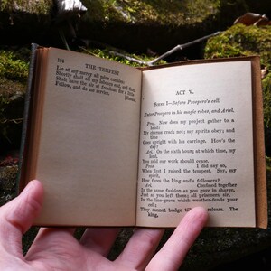 May include: An open, antique book with aged, yellowed pages. The text on the right page reads "ACT V. Scene 1 - Before Prospero's cell." The book is held open, revealing the text of Shakespeare's "The Tempest."