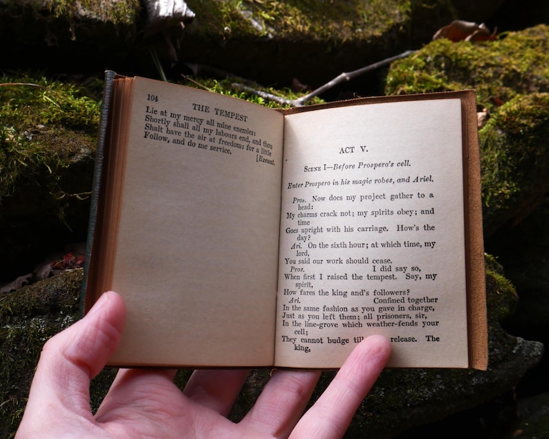 May include: An open, antique book with aged, yellowed pages. The text on the right page reads "ACT V. Scene 1 - Before Prospero's cell." The book is held open, revealing the text of Shakespeare's "The Tempest."