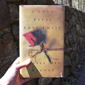 Może przedstawiać: Książka zatytułowana "A Long Fatal Love Chase" autorstwa Louisy May Alcott. Okładka przedstawia czerwoną różę zawiązaną sznurkiem.
