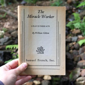 Op de afbeelding: Een beige paperbackboek getiteld The Miracle Worker, A Play in Three Acts door William Gibson, uitgegeven door Samuel French, Inc. De kaft bevat zwarte tekst, een kleine illustratie van een wapenschild en dunne zwarte verticale lijnen.