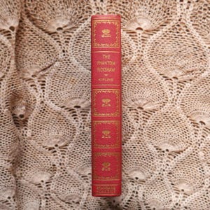 May include: A red hardcover book titled "The Phantom Rickshaw" by Kipling. The spine features gold lettering and decorative gold accents. The book is part of the Standard Classics series.