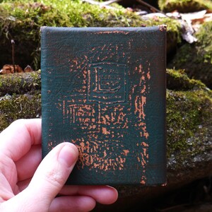 May include: A small, rectangular, dark green book with a textured cover and embossed design. The cover shows signs of wear with orange-brown accents. The book is held in a hand against a mossy background.